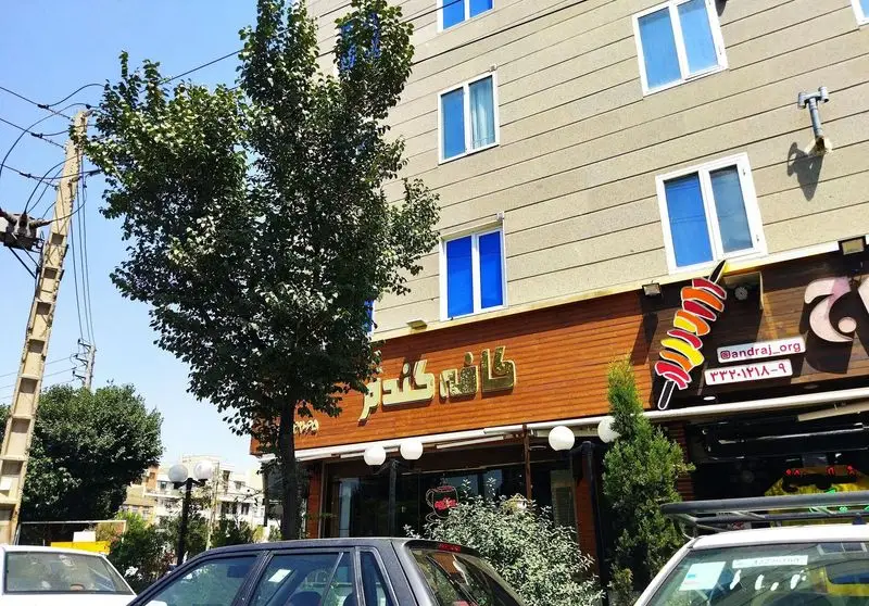 نمای بیرونی کافه گندم. تابلوی طلایی این کافه بر روی نمای چوبی ساختمان، همراه با پنجره‌های آبی و درختان سرسبز، زیر آسمان صاف.