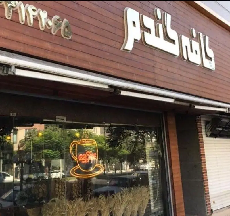 نمای بیرونی و سردر چوبی کافه گندم با تابلوی برجسته. نئون طرح فنجان قهوه در پنجره این کافی شاپ محیطی دلنشین را نشان می دهد.