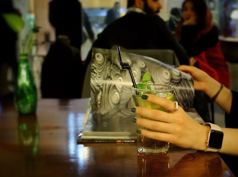 نوشیدن موهیتو در کافه مان و رو، مشتری در حال ورق زدن کتابی با طرح منحصر به فرد در محیط دنج کافه با میز چوبی.