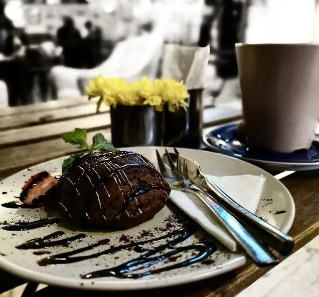 دسر شکلاتی بی‌نظیر کافه مان و رو با تزئین سس شکلات، توت فرنگی و نعنا، کنار قهوه و گل‌های زرد در محیط آرام کافه