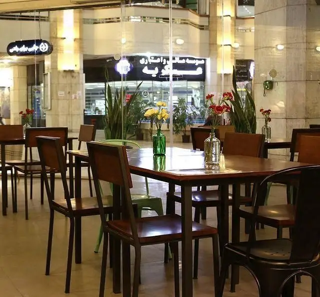 فضای داخلی دنج کافه مان و رو با میز و صندلی‌های چوبی، گلدان‌های گل زیبا و نورپردازی گرم، محیطی عالی برای استراحت.