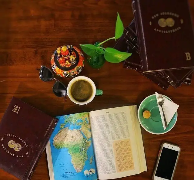 میز چوبی کافه مان و رو با یک فنجان قهوه، کتاب‌های دایره المعارف، نقشه‌ آفریقا، عینک و موبایل. فضای دنج و آرام برای مطالعه و لذت بردن از نوشیدنی.