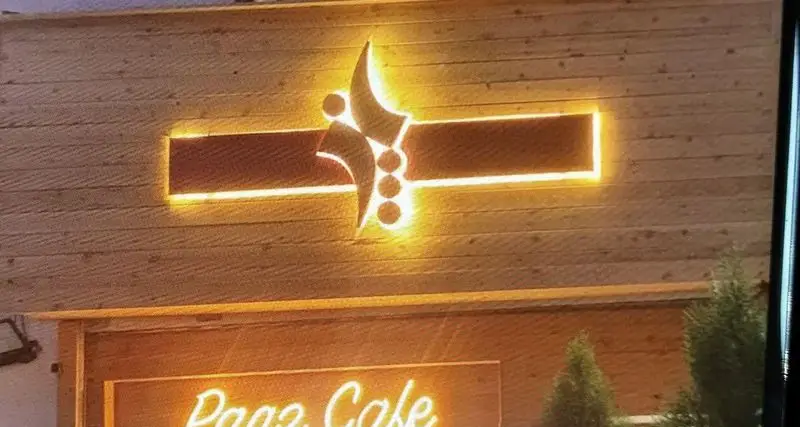 نمای بیرونی جذاب و دلنشین کافه پاز با دکوراسیون چوبی مدرن، لوگوی نورانی منحصر به فرد و تابلوی نئونی "Paz Cafe"، ایده‌آل برای اوقات خوش.