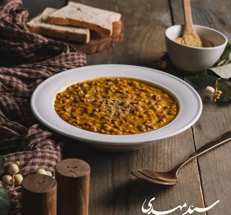 تصویری دلنشین از آش یا عدسی گرم و خوشمزه سید مهدی، غذای سنتی و مقوی با ادویه‌جات معطر، سرو شده با نان تازه روی میز چوبی.