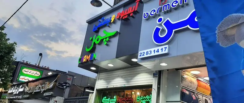 نمای بیرونی آبمیوه و بستنی چمن با تابلوهای نئون سبز و آبی، به همراه نام انگلیسی Chaman و شماره تماس 22831417.