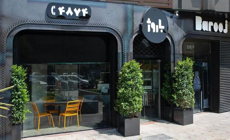 نمای بیرونی فست فود باروژ با معماری مدرن و تابلوی درخشان Barooj، شامل علامت ۲۴۷۴۴ و صندلی‌های نارنجی رنگ در فضای داخلی.