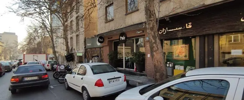 نمای بیرونی فست فود باروژ در یک روز آفتابی در خیابانی پرتردد در تهران با خودروهای پارک شده و موتورسیکلت‌های آماده تحویل غذا.