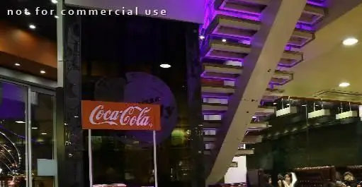 فضای داخلی شیک فست فود شایلی با پله‌های مدرن نورپردازی شده بنفش، تبلیغ کوکاکولا و مشتریان در حال لذت بردن از غذا.