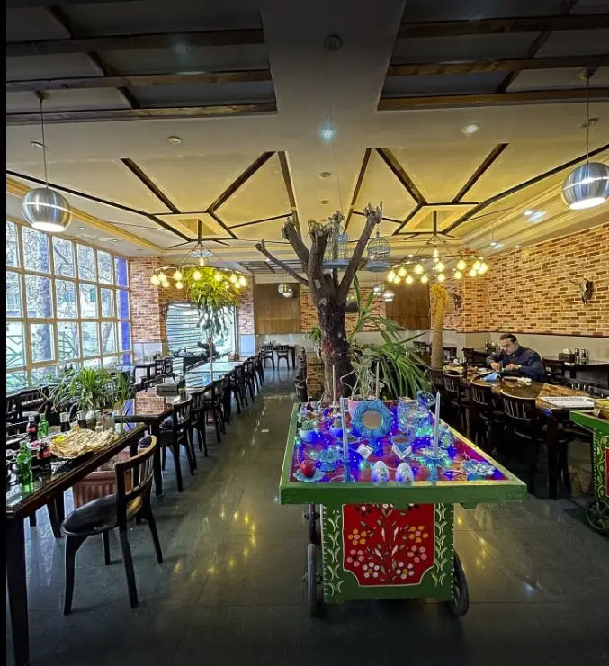 فضای داخلی طباخی کاج: رستورانی با دیوارهای آجری، پنجره‌های بزرگ، سقف طرح‌دار، درختان داخلی و دکوراسیون گرم و سنتی.