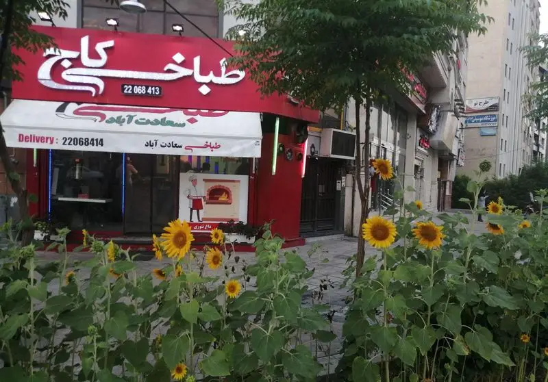 تصویری از طباخی کاج در منطقه سعادت آباد تهران، با تابلوی قرمز و سفید، شماره‌های تماس و ردیفی از گل‌های آفتابگردان زیبا.