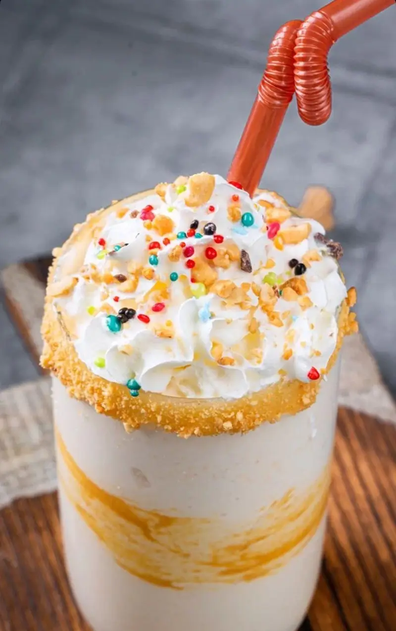 میلکشیک وانیلی خوشمزه کافه بیلیارد پلن بی با خامه فراوان، اسپرینکل‌های رنگی شاد و تزیین آجیل‌های تازه. بهترین انتخاب برای دسر و نوشیدنی خنک و دلپذیر.