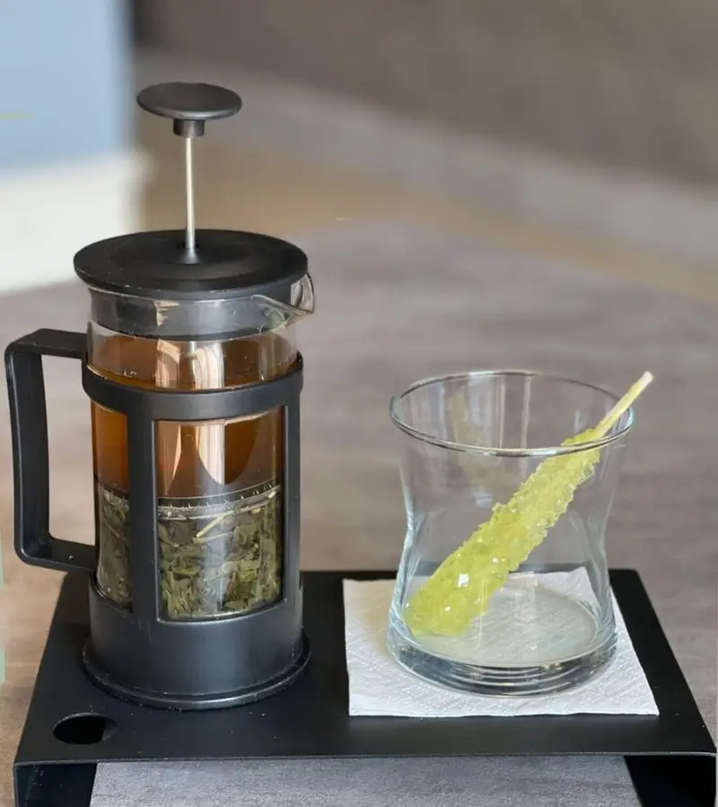 تصویری از فرنچ پرس چای سبز با برگ‌های چای در حال دم کشیدن، کنار یک لیوان با نبات شاخه‌ای سبز روی سینی مشکی، آماده سرو در کافه بیلیارد پلن بی.