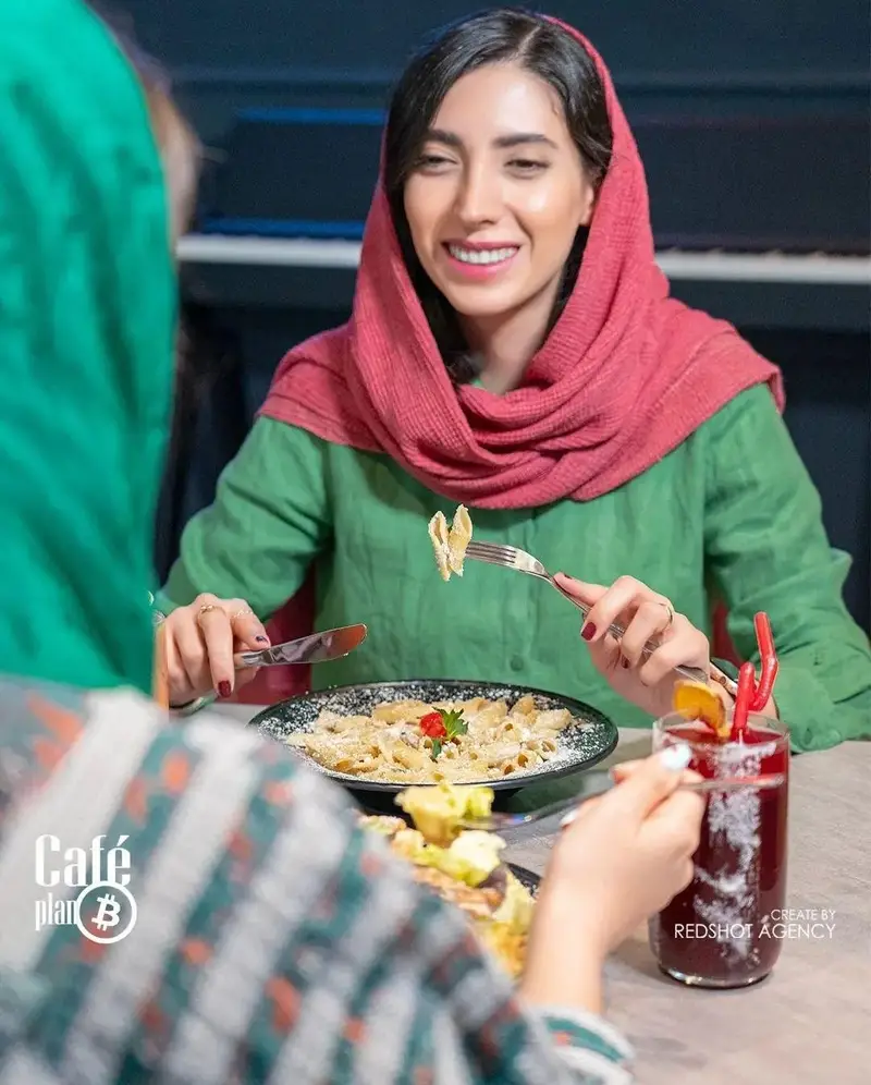 تصویری از دو خانم در کافه پلن بی، لذت‌بردن از پاستا و نوشیدنی‌های دلپذیر. زن با روسری قرمز لبخندزنان پاستا میل می‌کند.