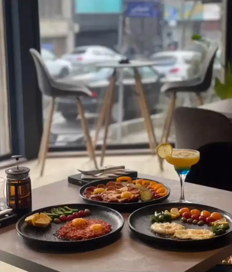 نمای جذاب از فضای داخلی کافه بیلیارد پلن بی، شامل میزهای چوبی، صندلی‌های شیک، انواع صبحانه و نوشیدنی‌های رنگارنگ با چشم‌انداز خیابان.