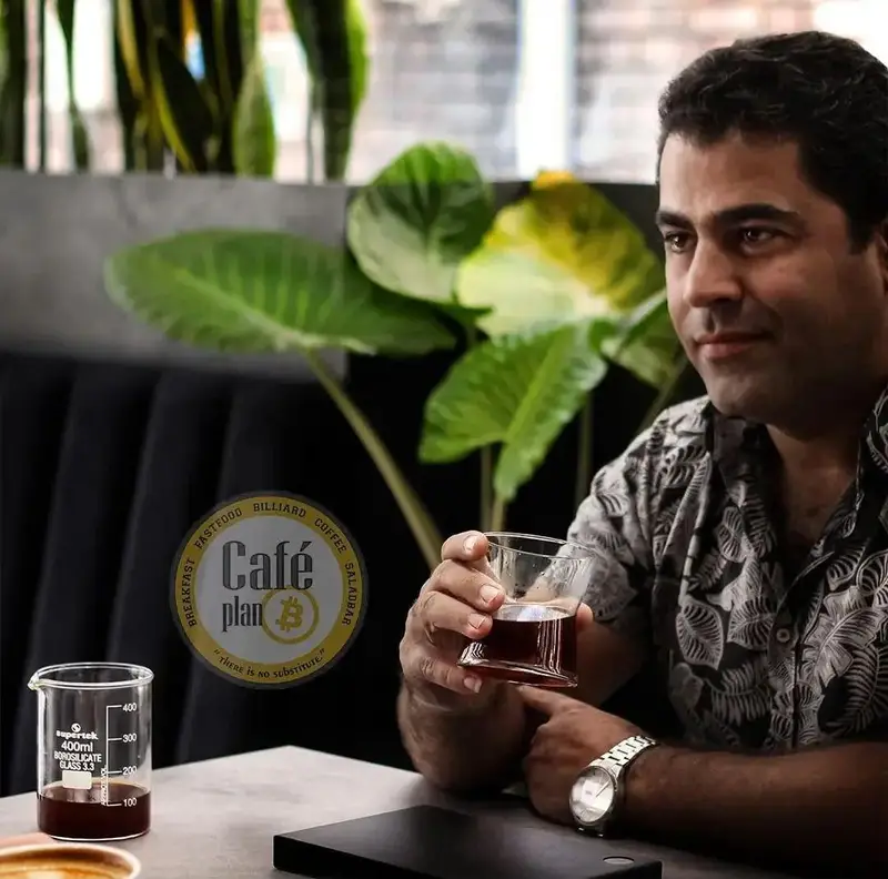 مردی خوشحال در حال نوشیدن قهوه در کافه بیلیارد پلن بی. لذت بردن از فضای داخلی مدرن و آرام با منوی متنوع صبحانه، فست فود و سالاد بار.