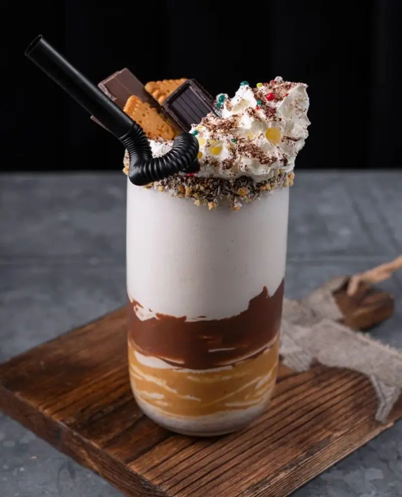 میلک شیک سه‌لایه ویژه کافه بیلیارد پلن بی، شامل شکلات، کارامل و وانیل، با خامه فرم گرفته، شکلات و بیسکویت تزیین شده است.