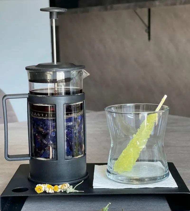 دمنوش خاص چای پروانه آبی در فرنچ پرس، با نبات شاخه ای سبز در لیوان شیک، سرو شده در فضای کافه بیلیارد پلن بی.