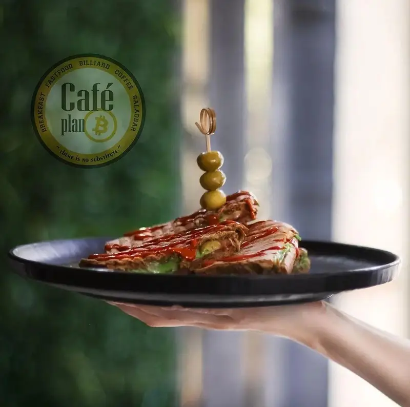 ساندویچ فست فود کافه بیلیارد پلن بی با تزئین زیتون و سس قرمز روی بشقاب مشکی. این کافه، صبحانه، قهوه و سالادبار دارد.