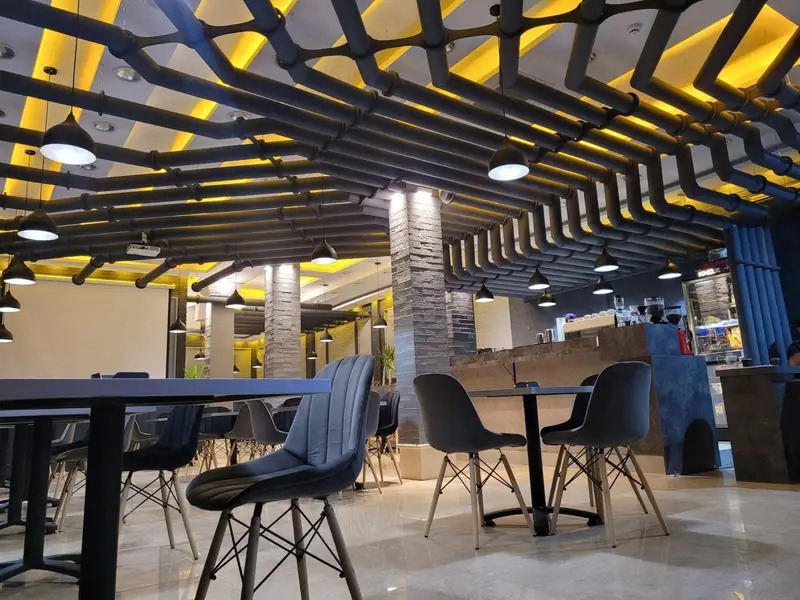 تصویری از طراحی داخلی منحصر به فرد کافه بیلیارد پلن بی: سقف صنعتی با لوله‌های تیره، نورپردازی گرم، و چیدمان صندلی‌های مدرن برای راحتی مشتریان.