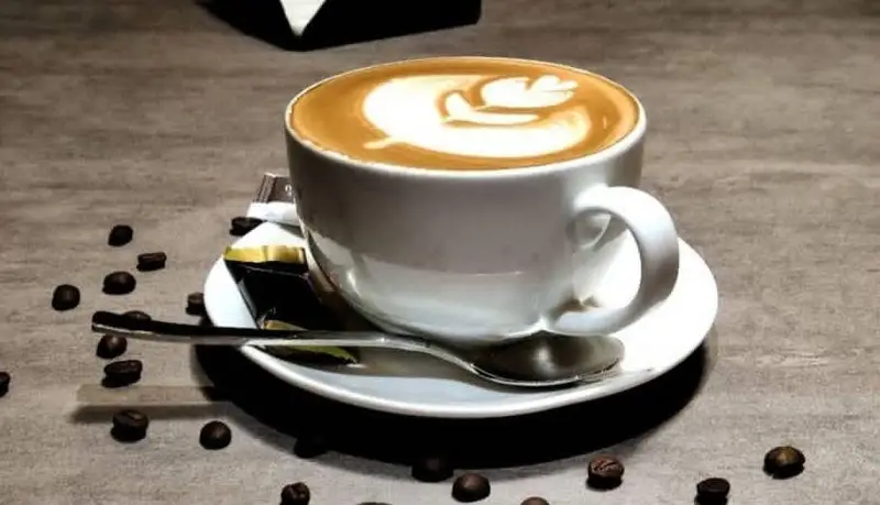 یک فنجان کاپوچینو با طرح لاته آرت دلنشین، به همراه دانه های قهوه در کافه بیلیارد پلن بی. بهترین انتخاب برای یک نوشیدنی آرام.