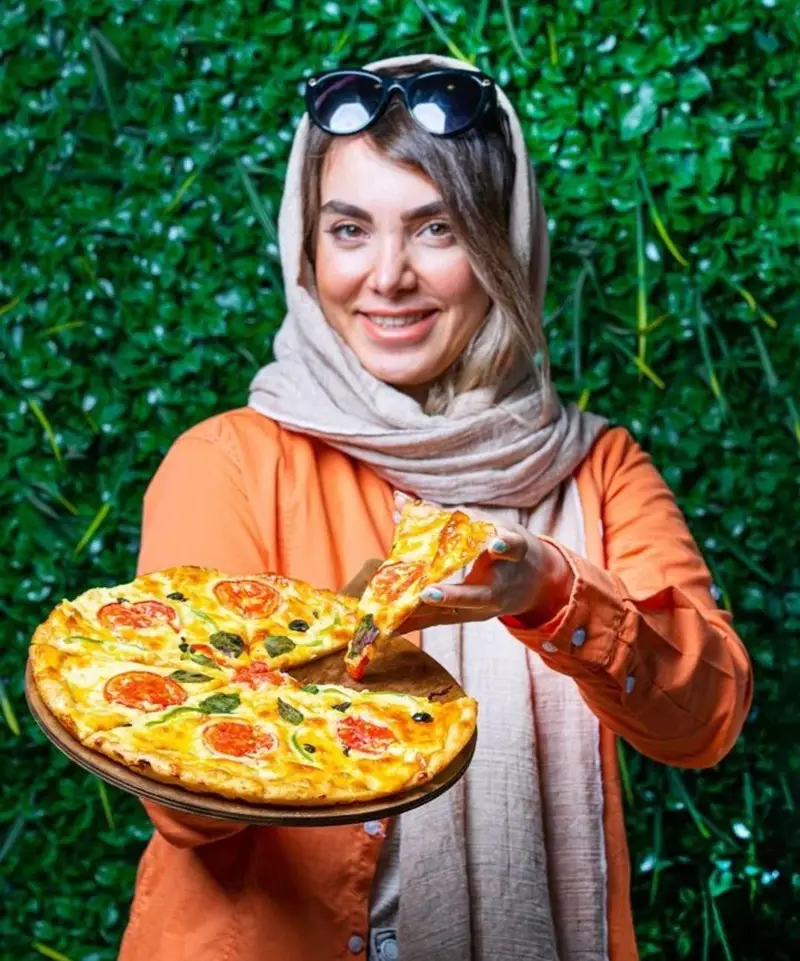 تصویری دلنشین از یک زن خندان در کافه بیلیارد پلن بی که پیتزای تازه و لذیذ را با عشق تقدیم می کند. مکانی عالی برای غذا و تفریح.