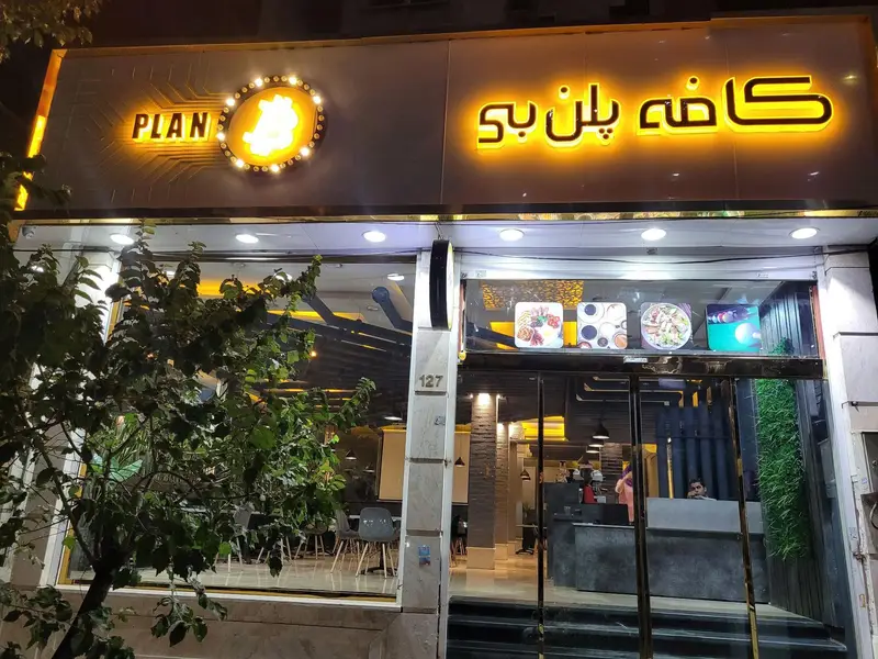 نمای ورودی شبانه کافه و بیلیارد پلن بی با تابلوی روشن، لوگوی بیت‌کوین و ویترین غذا، مکانی برای استراحت و تفریح.