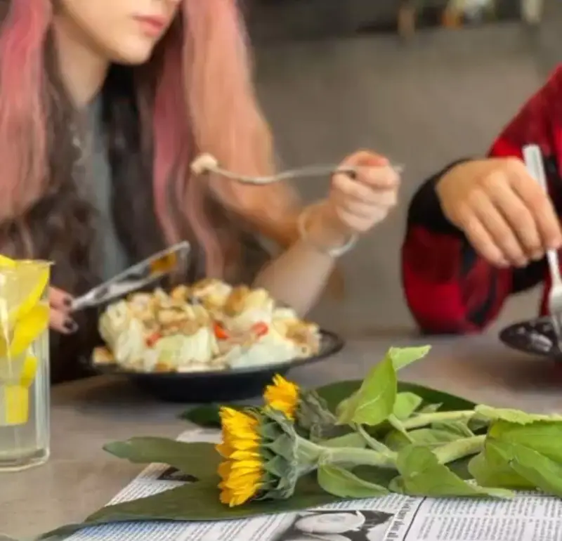 تجربه غذای خوشمزه و لحظات دلنشین در کافه بیلیارد پلن بی. دو دوست در حال صرف ناهار یا شام، نوشیدنی تازه و گل آفتابگردان روی میز.