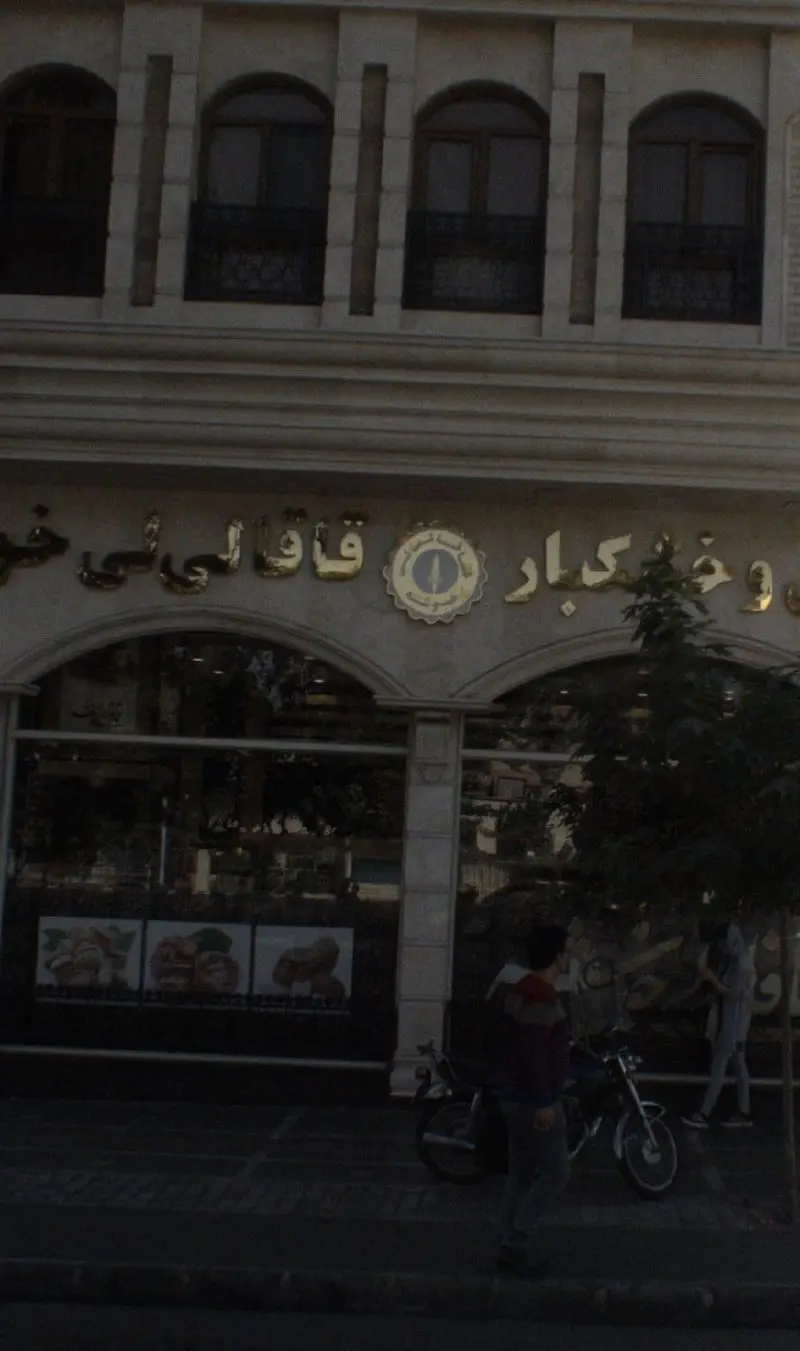 تصویر نمای بیرونی قنادی قاقالی لی با تابلو برجسته طلایی، ورودی شیک، و ویترین نمایش شیرینی و کیک‌های تازه در مرکز شهر.
