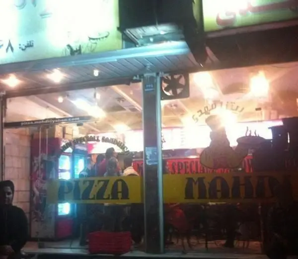 نمای بیرونی و پرنور فست فود پیتزا مهدی در شب با تابلوهای بزرگ “PIZZA” و “MAHDI” روی شیشه ویترین و آدرس ایمیل pizza.mahdi@yahoo.com.