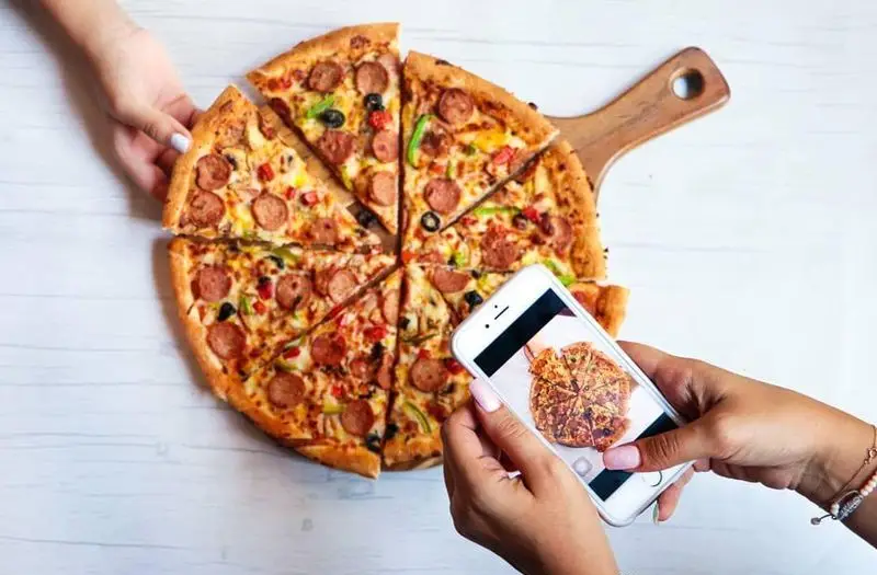 عکس هوایی از پیتزای سوسیس، فلفل دلمه‌ای و زیتون فست فود شیلا؛ یک دست در حال برداشتن اسلایس و دست دیگر با گوشی از پیتزا عکس می‌گیرد.