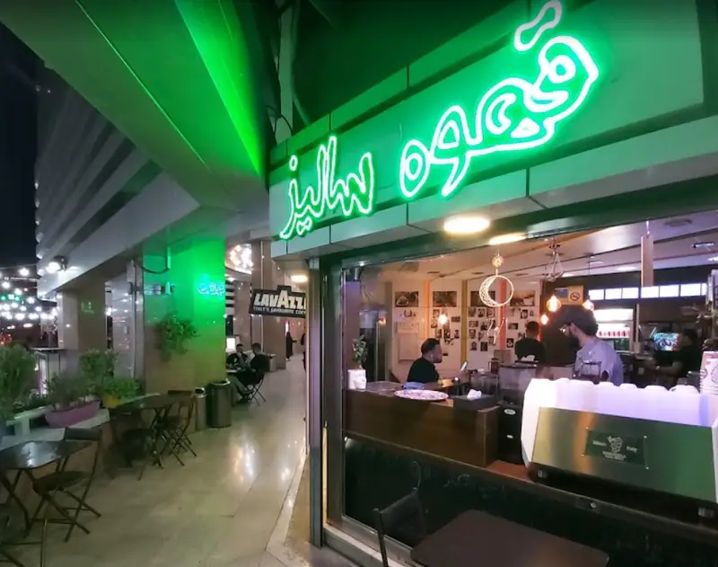 نمای شب کافه سالیز با تابلوی نئون درخشان "قهوه سالیز"، مشتریان در داخل و میز و صندلی‌های فضای باز.