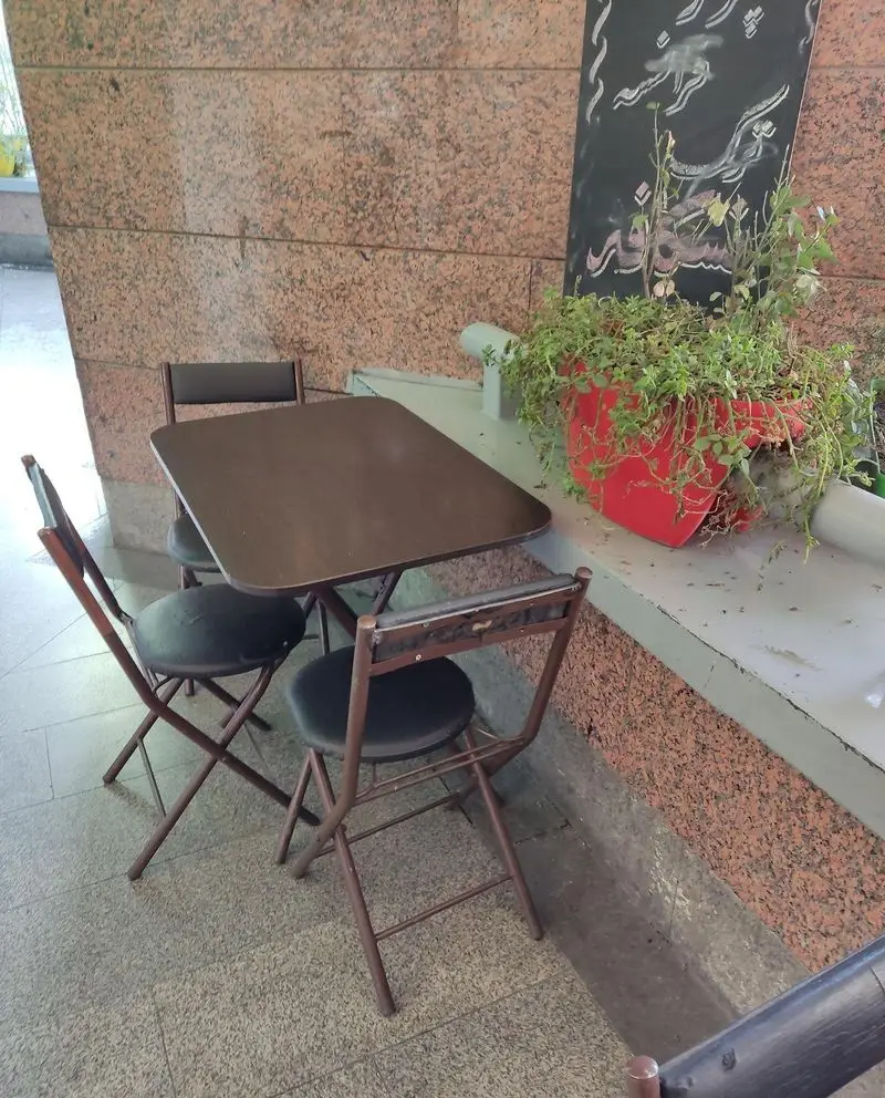 فضای کافه سالیز با میز و صندلی‌های قهوه‌ای تاشو، گلدان قرمز گیاهان آویز و تخته سیاه تزئینی. محیطی مناسب برای استراحت و گپ‌وگفت.