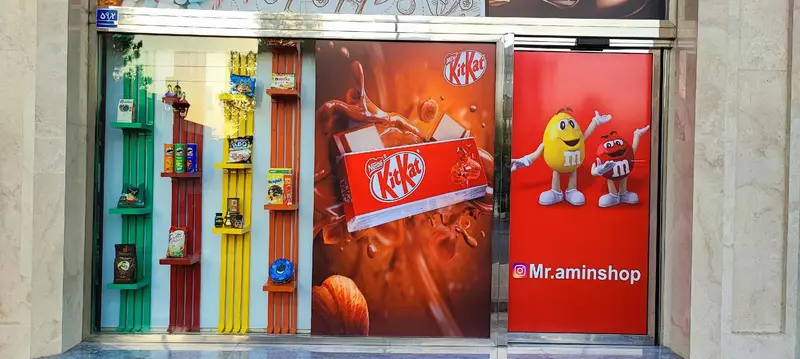 ویترین چشم‌نواز فروشگاه مستر امین شاپ با قفسه‌های رنگی سبز، قرمز، زرد پر از تنقلات، قهوه و چیپس. پوستر کیت کت و M&M's با آدرس اینستاگرام @Mr.aminshop.