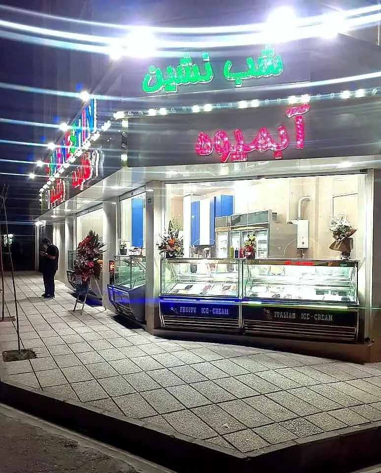 نمای شبانه ویتامینه و فالوده شب نشین، با تابلوهای نئونی درخشان آبمیوه، ویترین بستنی میوه‌ای و بستنی ایتالیایی روشن.