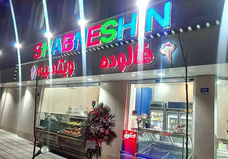 نمای خارجی مغازه ویتامینه و فالوده شب نشین در شب، با تابلوهای رنگارنگ، عرضه آبمیوه، ویتامینه، فالوده و بستنی.