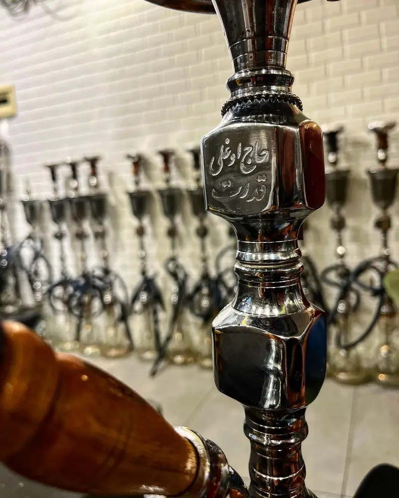 تصویر نزدیک از تنه قلیان فلزی و براق با حکاکی برند معروف حاج اوغلی قدرت در کافه عربی میر مهنا، نمادی از قلیان‌های اصیل و باکیفیت.