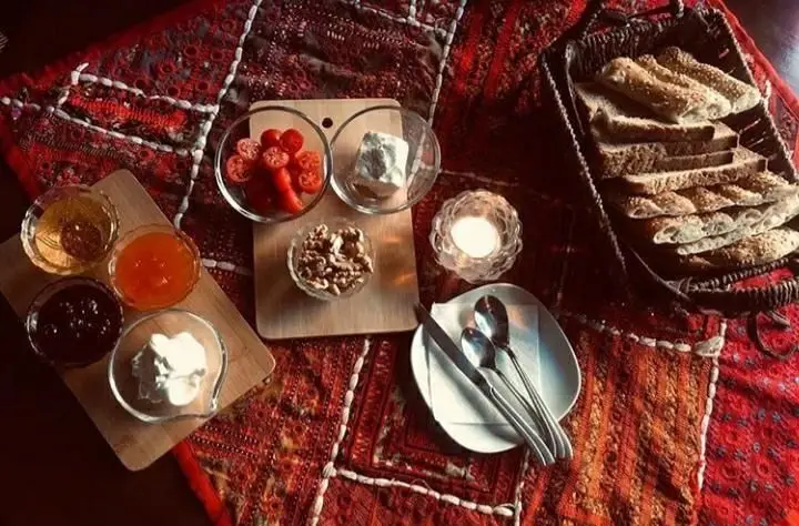 میز صبحانه کامل و دلچسب کافه لدی با نان‌های تازه، پنیر محلی، گوجه، گردو، عسل و چندین نوع مربا و خامه در فضایی گرم و سنتی.