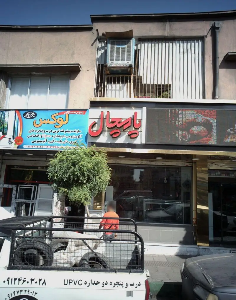 نمای خارجی شیرینی فروشی پامچال با تابلوی زیبا و ویترین شیشه‌ای که دسرها را به نمایش گذاشته، در کنار یک مرکز پنجره‌سازی.