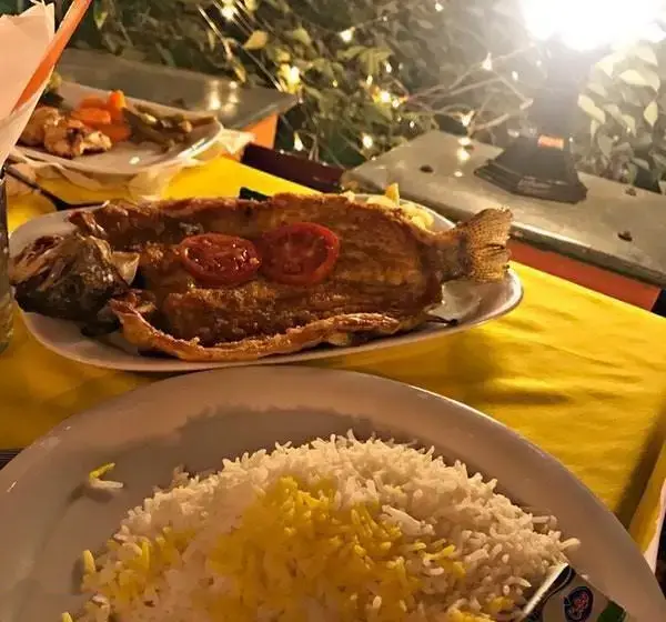 شام لذیذ در رستوران باغ بهشت: ماهی سرخ شده طلایی با برنج زعفرانی و مخلفات کنار میز در فضایی آرام و سرسبز با نورپردازی دلنشین.