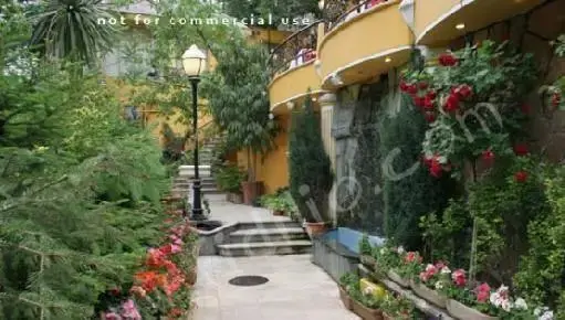 نمایی زیبا از باغ رستوران باغ بهشت با مسیر سرسبز، گل‌های رنگارنگ، آبشار کوچک و ساختمان زرد رنگ با بالکن‌های دلنشین.