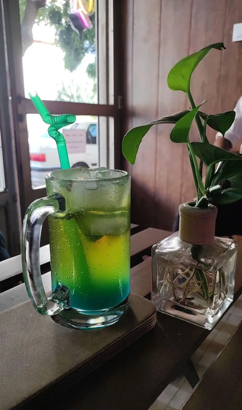 یک نوشیدنی رنگارنگ و با طراوت در لیوان دسته دار امیر کافه، همراه با نی سبز فانتزی و گلدان کوچک تزئینی کنار پنجره.
