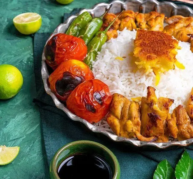 یک بشقاب چلو جوجه کباب شمرون، با ته دیگ زعفرانی، گوجه و فلفل کبابی، همراه با لیمو ترش و سس. غذای اصیل ایرانی.