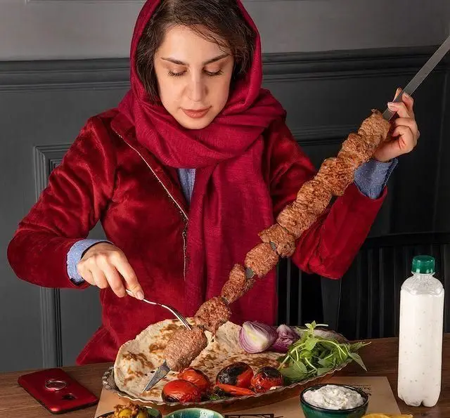 زن با حجاب قرمز در رستوران شمرون کباب، در حال میل کباب کوبیده با نان، گوجه، سبزی و دوغ. لذت غذای اصیل ایرانی.