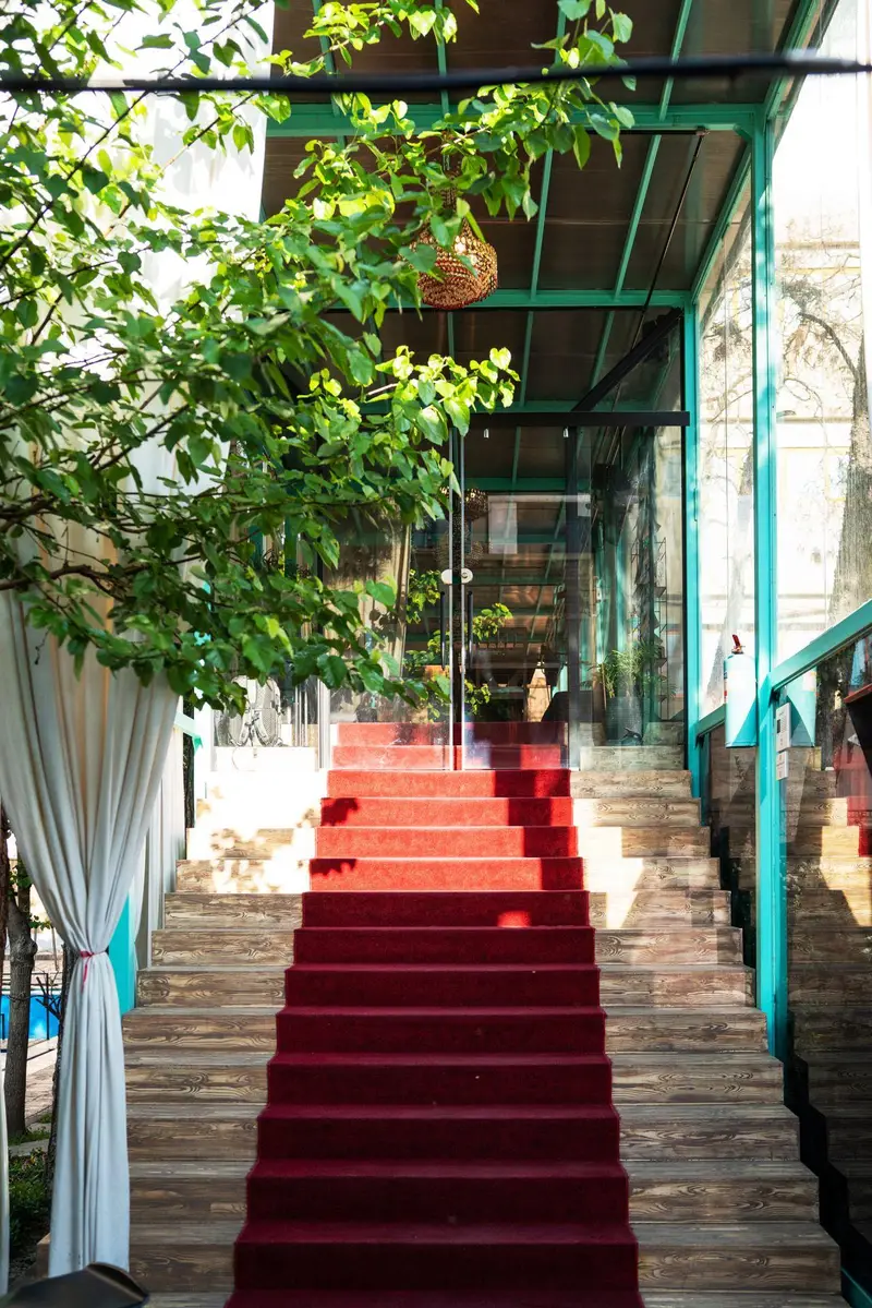 ورودی چشمگیر کافه‌حیات هزار افسان با پله‌های قرمز مخمل، دیوارهای شیشه‌ای و سازه سبز، فضایی دنج و خاص برای مهمانان.