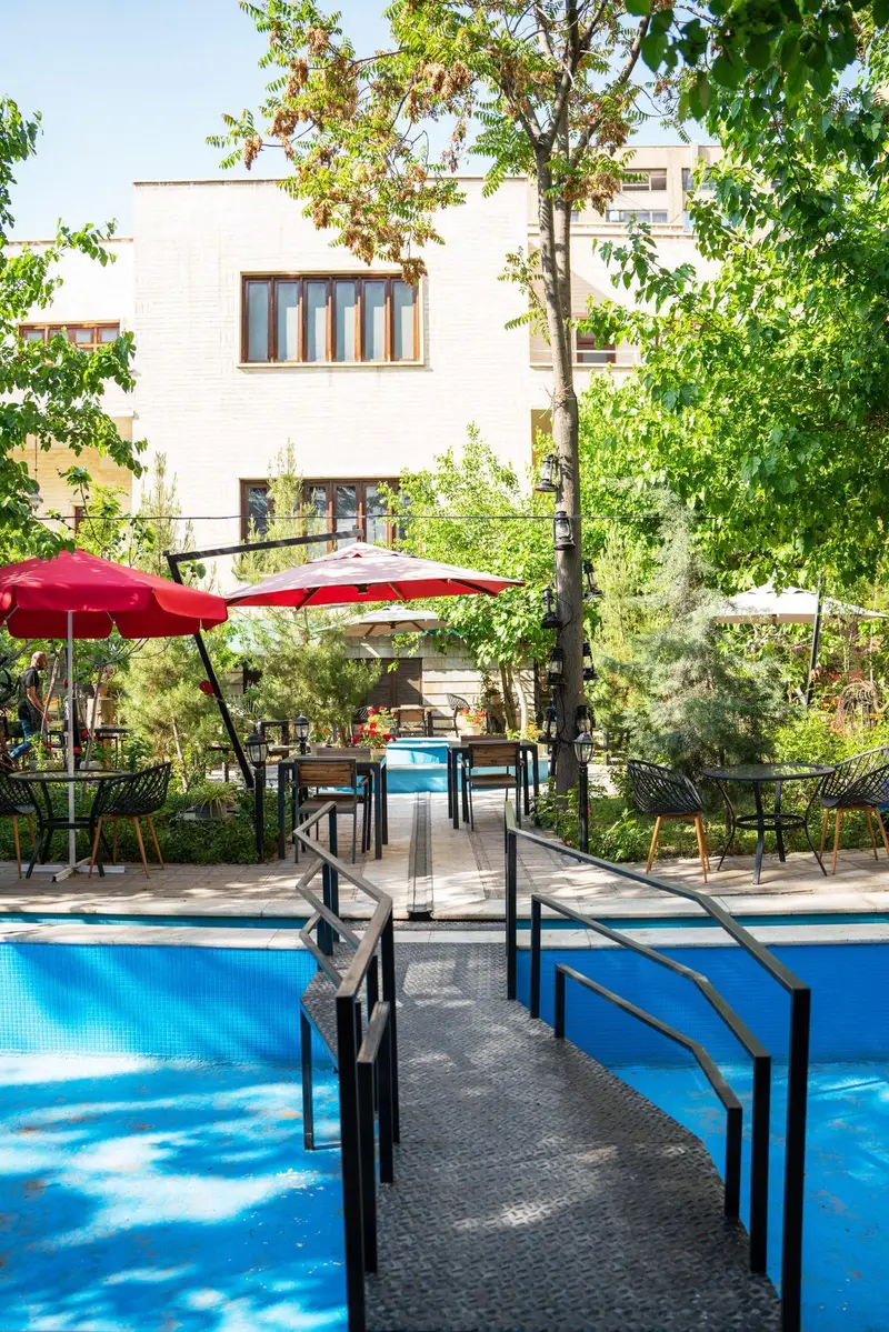 فضای بیرونی و دلنشین کافه‌حیات هزار افسانْ با میز و صندلی‌های چیده شده، چترهای قرمز، درختان سرسبز و آبنمای آبی. مناسب تابستان.