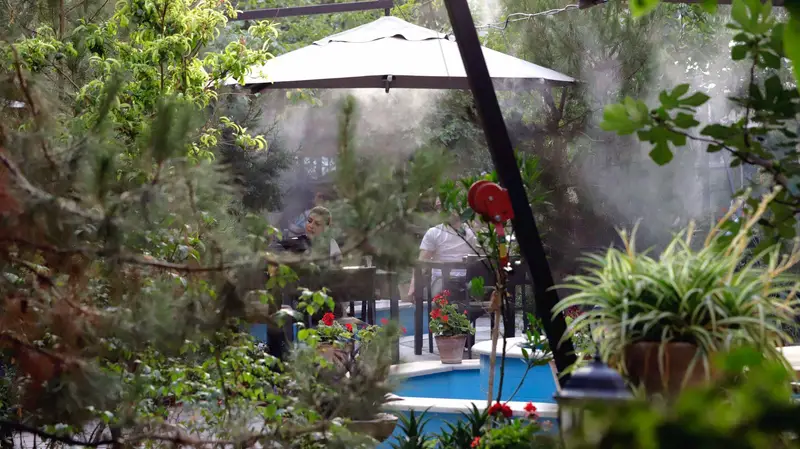 کافه‌حیات هزار افسانْ: فضای سبز و دلنشین کافه در فضای باز با مشتریان زیر مه خنک‌کننده و درختان سرسبز، مناسب برای تابستان.