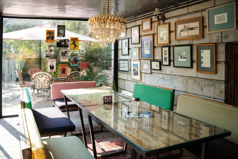 فضای دلنشین و شیک کافه‌حیات هزار افسانْ با میز بزرگ شیشه‌ای، چیدمان هنری دیوار و دید به باغ سرسبز بیرونی. محیطی آرام و زیبا برای مشتریان.