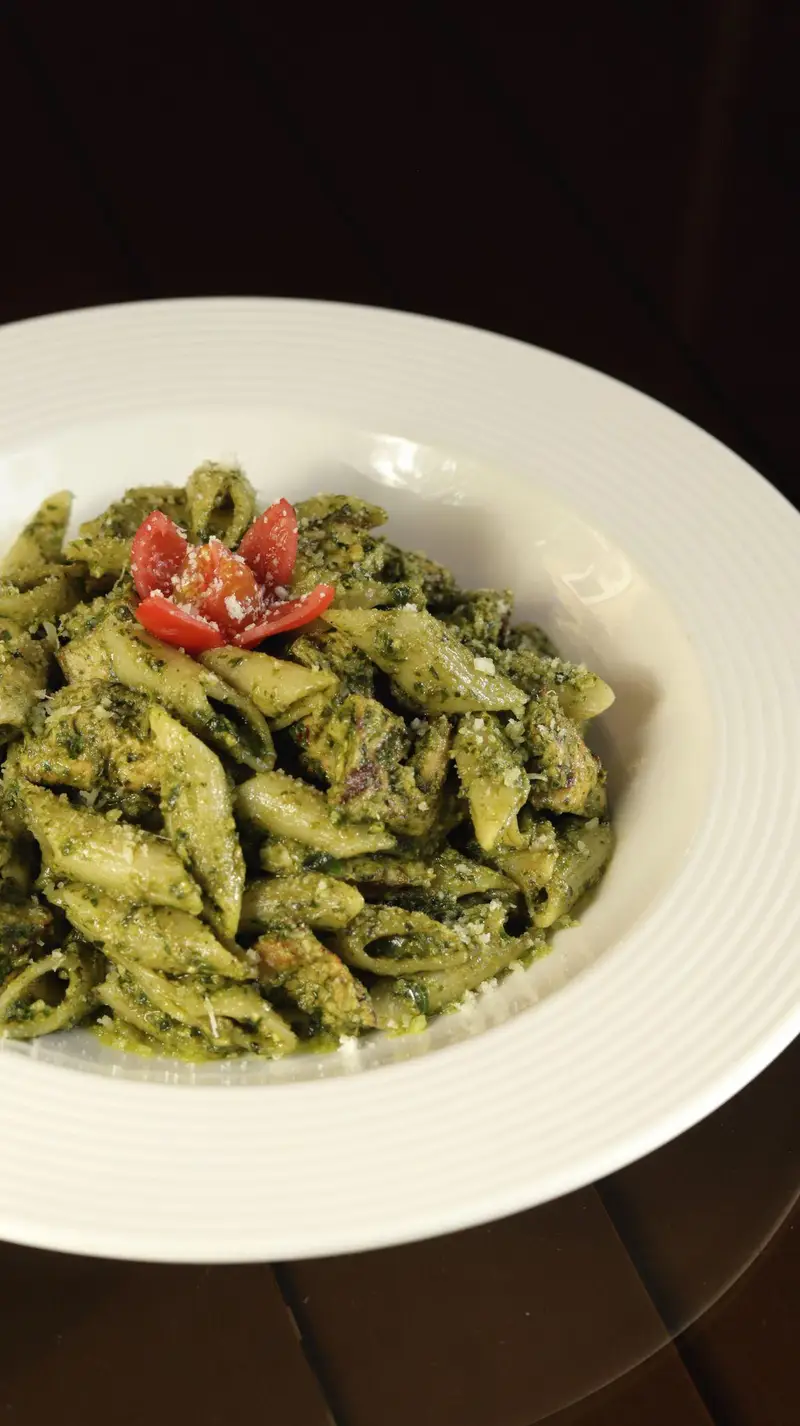 تصویر پاستا پنه پستو سبز جذاب و خوش‌رنگ با تکه‌های مرغ، پنیر رنده شده و تزیین گوجه، انتخابی دلپذیر از منوی کافه‌حیات هزار افسان.