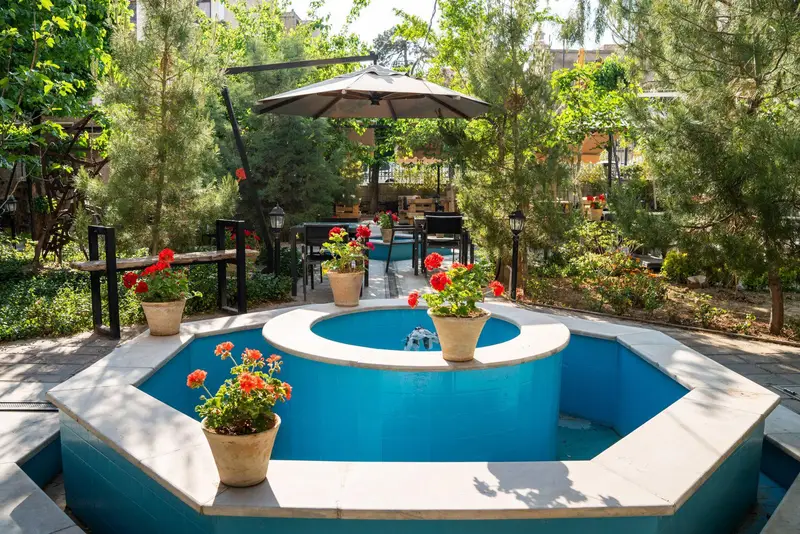 کافه‌حیات هزار افسانْ: فضای دلنشین و سرسبز با آبنمای فیروزه‌ای، میز و صندلی‌های چوبی در باغی آرام و زیبا مناسب دورهمی.