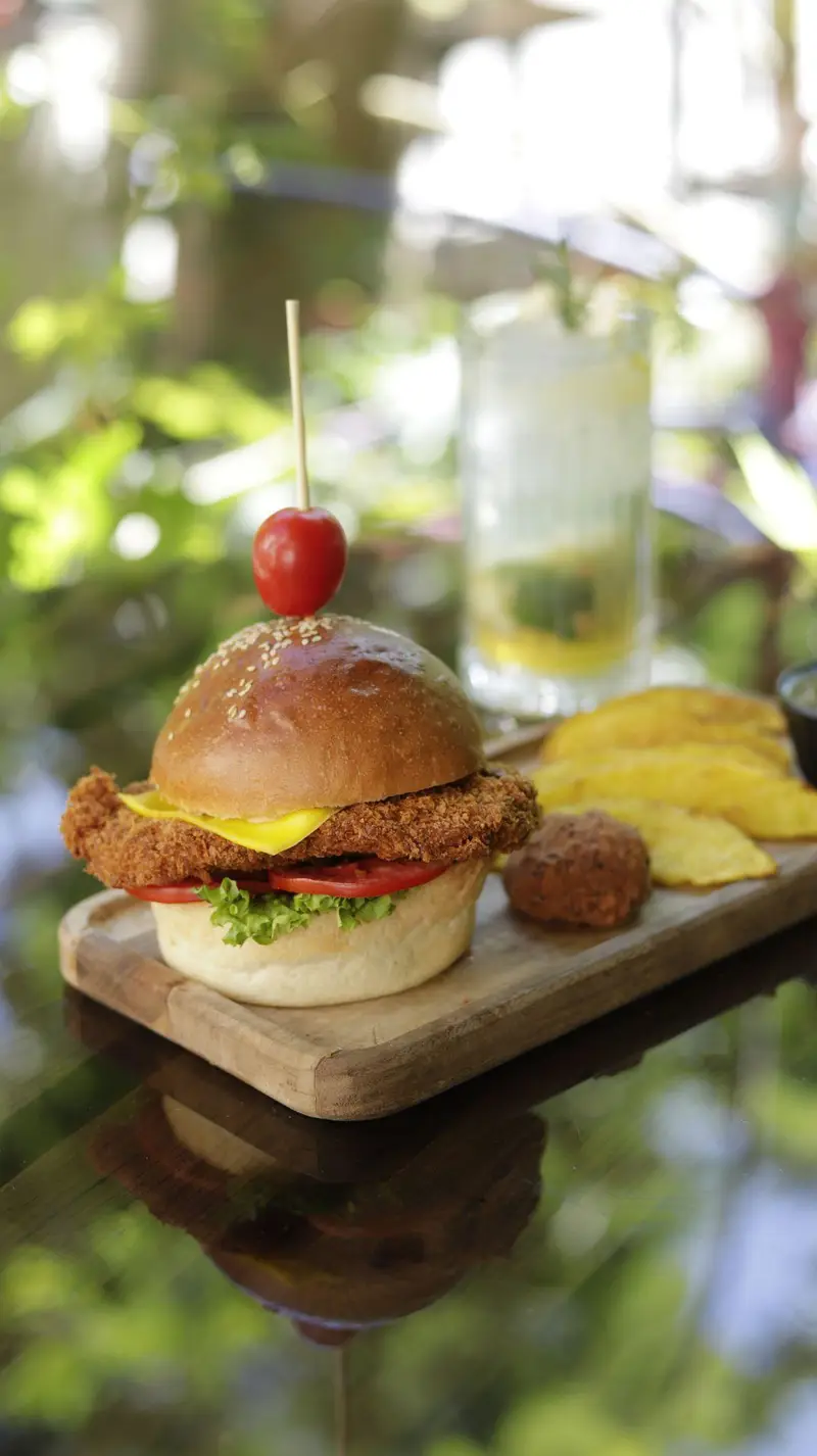 برگر شنیسل مرغ جذاب کافه‌حیات هزار افسانْ با پنیر، کاهو و گوجه، سرو شده با سیب‌زمینی و نوشیدنی در محیطی دلنشین.